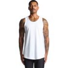 Man with tattoos wearing a white tank top and black pants, standing against a white background.