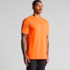 Man in an orange t-shirt and black pants standing against a plain gray background.