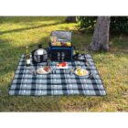 Picnic setup on plaid blanket with food, wine, glasses, and a cooler on grass near trees.