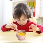 Young child in red shirt eating corn from a pink bowl with a spoon.