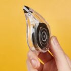 Hand holding a clear correction tape roller against an orange background.