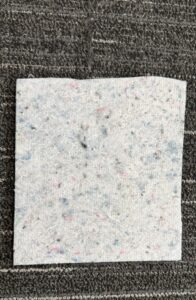 A recycled square of multicolored felt on a dark patterned carpet background.