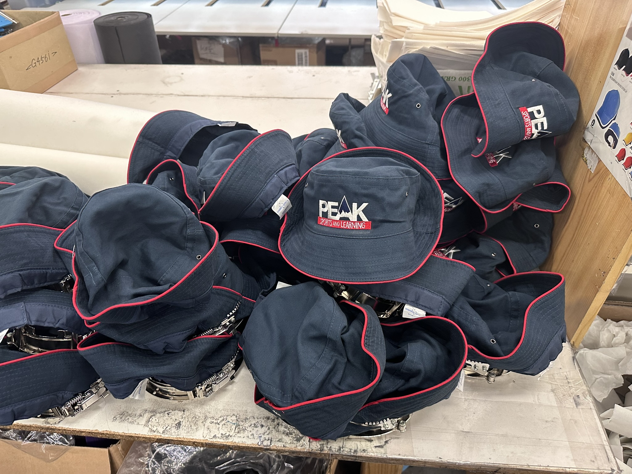 A pile of navy blue hats with red trim and embroidered logos sits on a table.