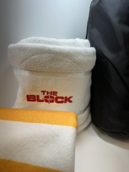 A white custom merch towel with THE BLOCK logo beside a yellow-striped towel and dark bag.