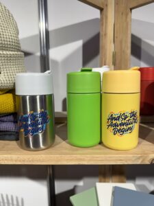 Three colorful thermoses, sponsored with And so the adventure begins, on a wooden shelf.
