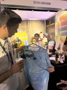 A man holds a blue tie-dye shirt at the APPA 2025 Buck Uniform booth with colorful apparel on display.