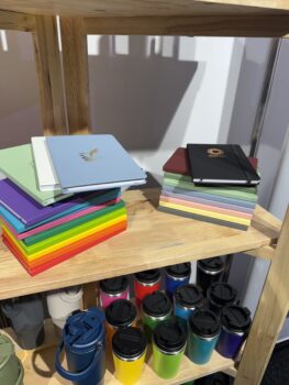 Colorful notebooks and reusable coffee cups for APPA 2025 displayed on wooden shelves.