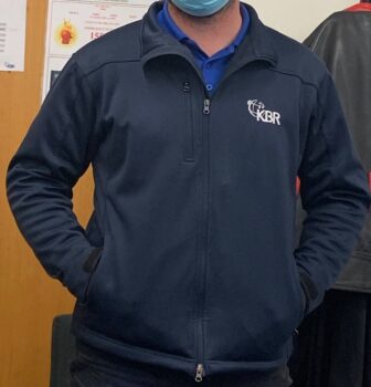 Person in a blue KBR jacket and face mask stands indoors, Embroider Logos proudly displayed.