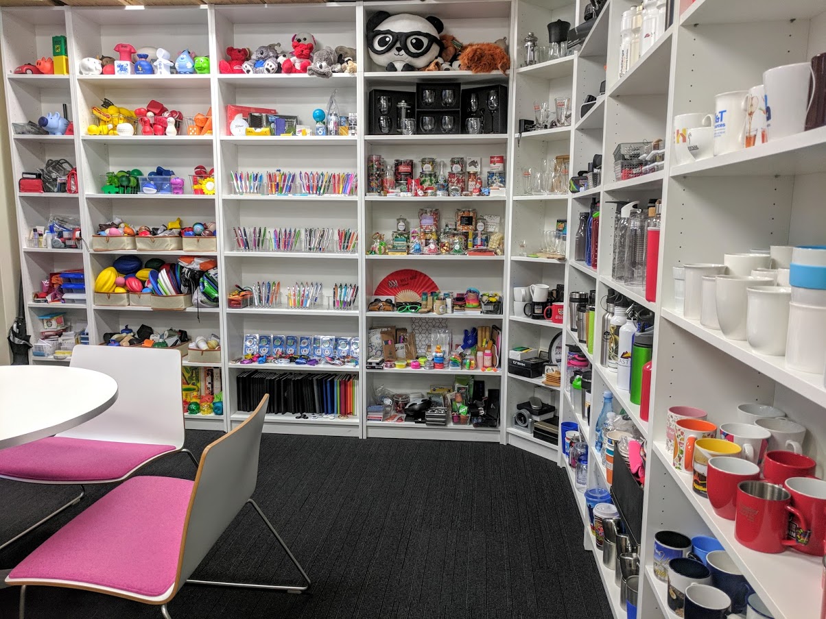 Shelves filled with colorful mugs, toys, and merchandise samples; a table with pink chairs.