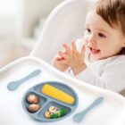 Smiling baby in a high chair with a divided plate of vegetables and utensils in front.