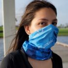 Person with long hair wearing a blue face covering, outdoors near water.