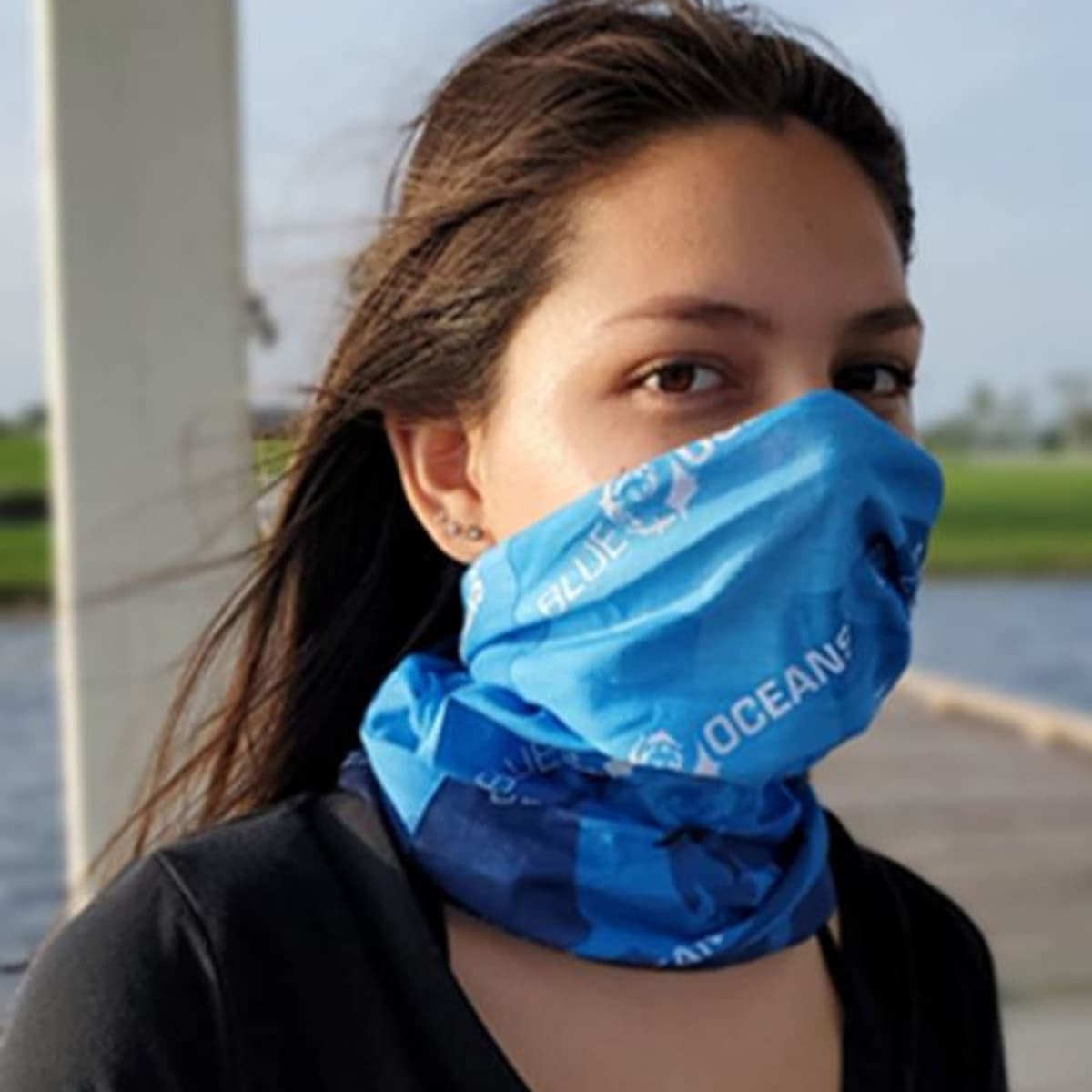 Person with long hair wearing a blue face covering, outdoors near water.