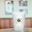 White cup with a green T-shirt and heart design, on a wooden table. Blurred background.