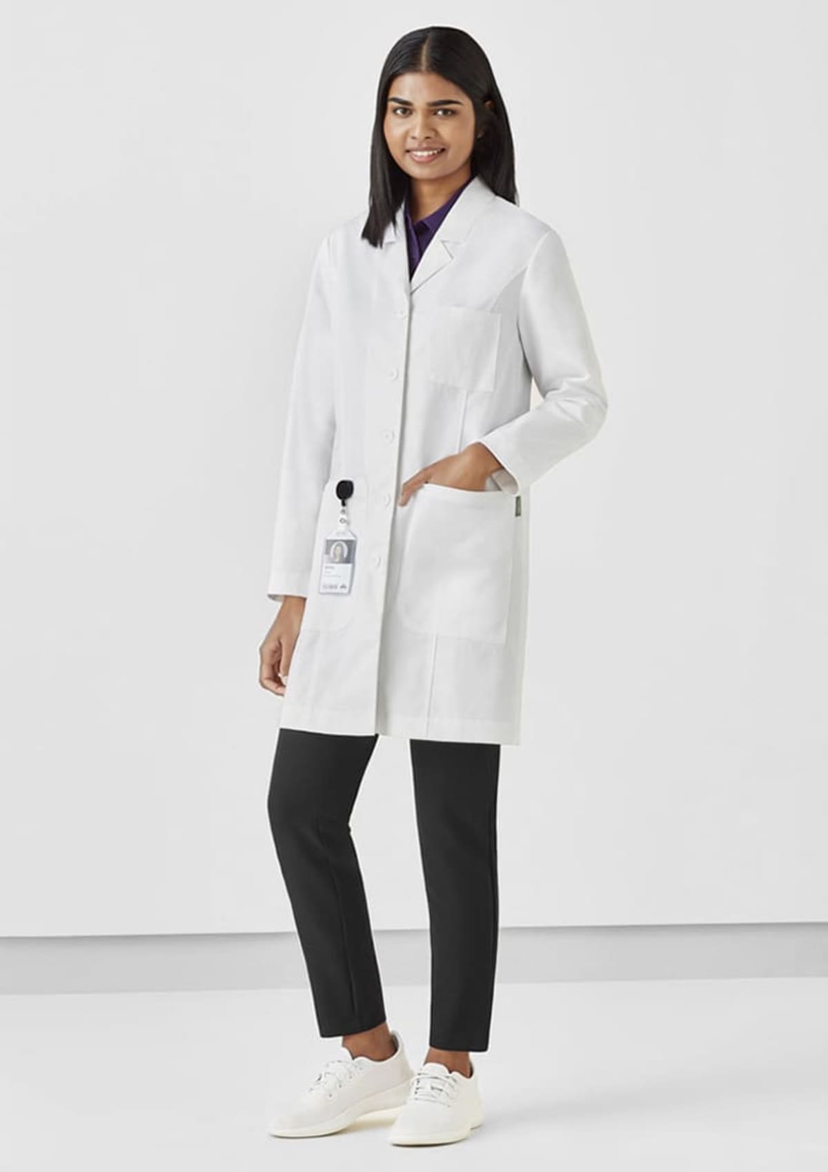 Person in a white lab coat and black pants smiling, standing in a neutral background.