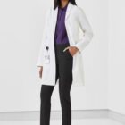 Person smiling, wearing a white lab coat, purple shirt, black pants, and white shoes.