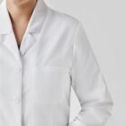 Person in a white lab coat with a pocket, smiling against a plain background.