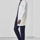 Person in a white lab coat and navy scrubs, standing and smiling in a minimalistic room.