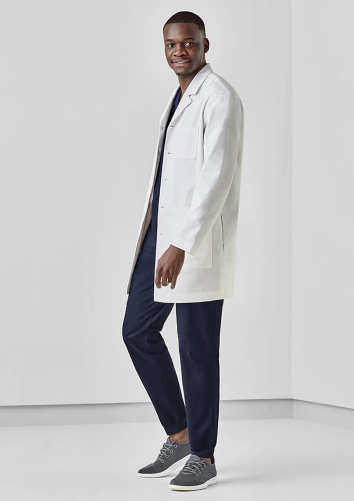Person in a white lab coat and navy scrubs, standing and smiling in a minimalistic room.