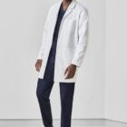 Person in white lab coat and dark scrubs standing and smiling in a plain room.
