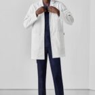 Person wearing a white lab coat over blue scrubs, standing and smiling.