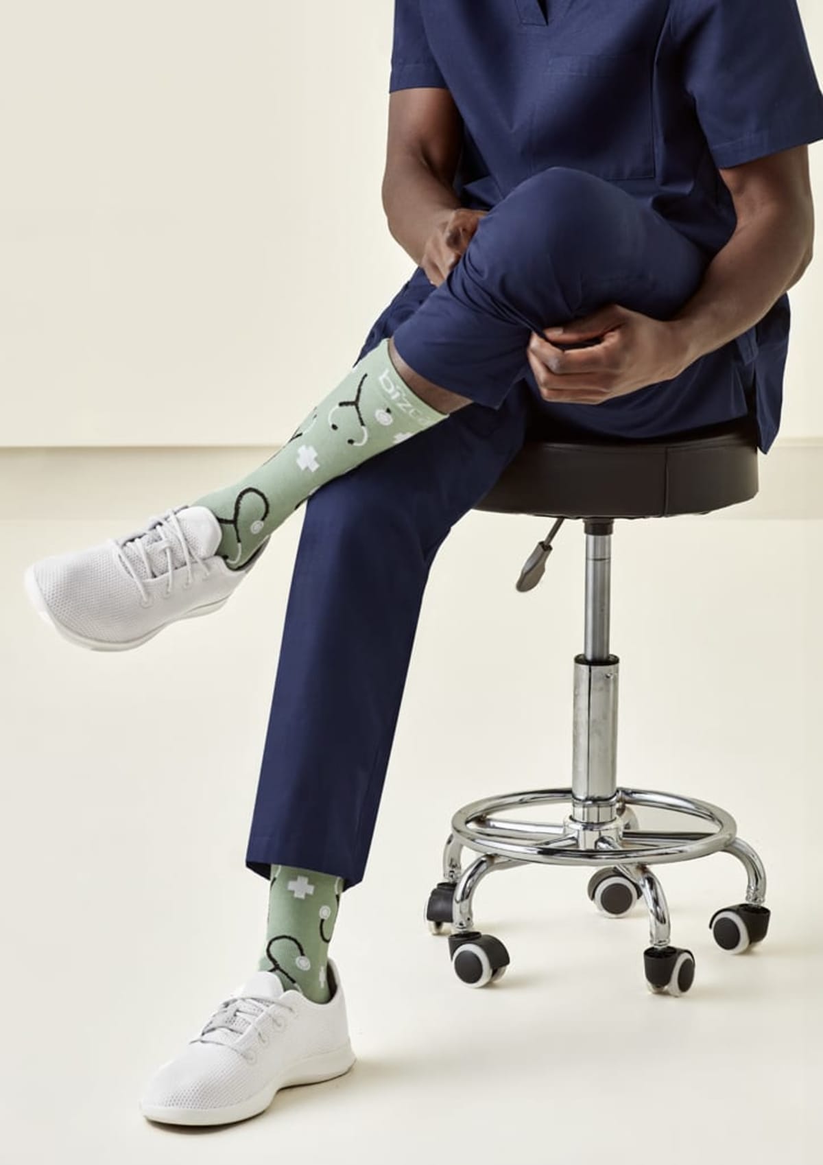 Person in scrubs sits on a stool wearing medical-themed socks and white sneakers.