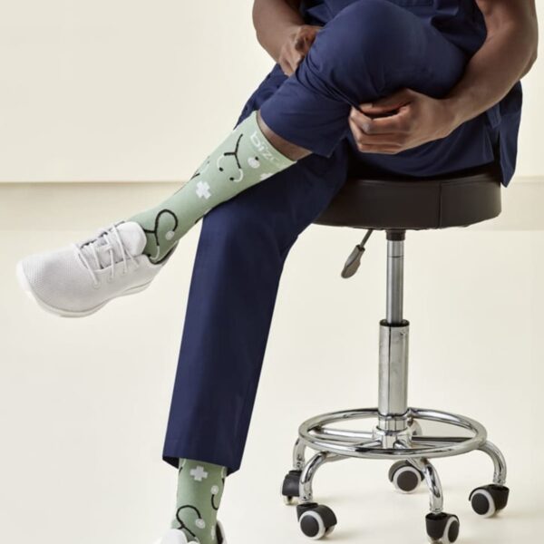 Person in scrubs seated on a stool, wearing green socks with medical symbols and white sneakers.