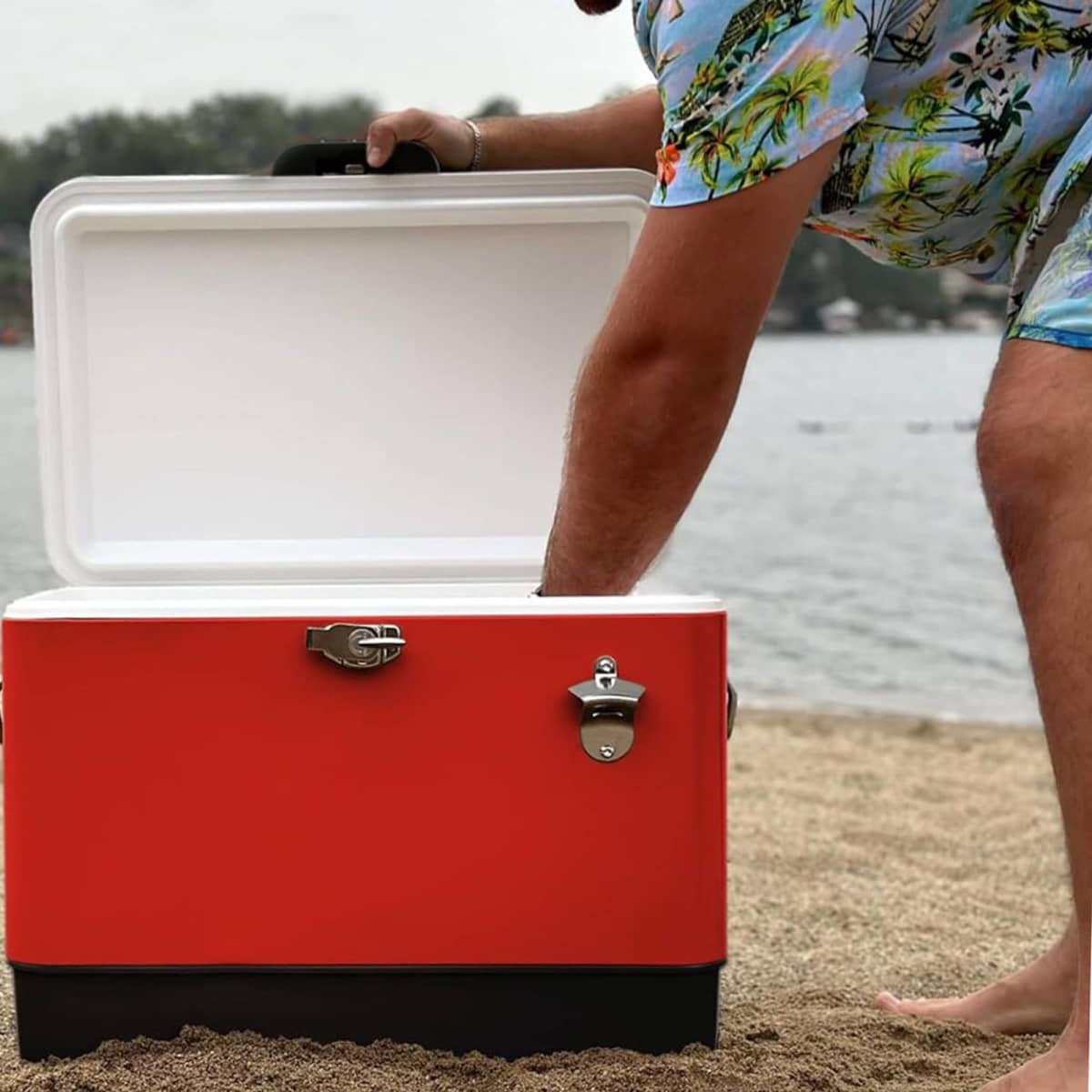Person in tropical shirt reaching into a red cooler on a sandy beach by the water.