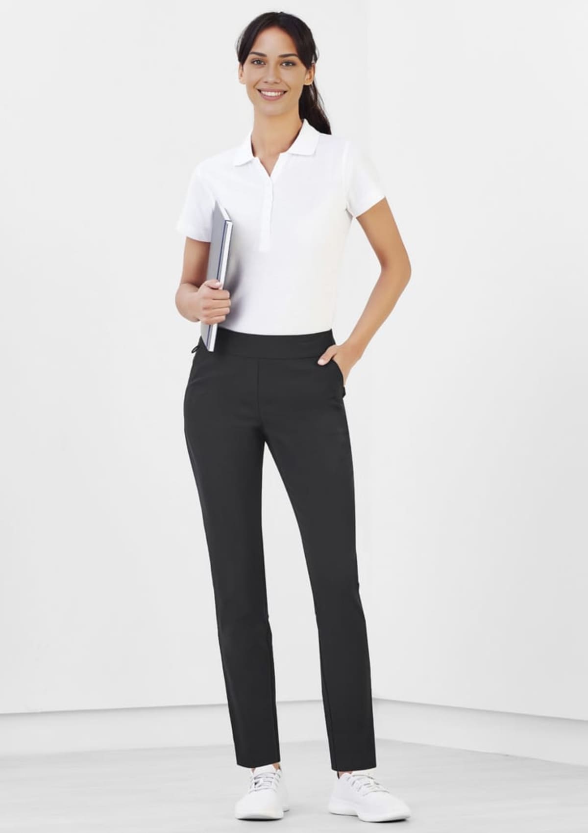 Smiling person in a white polo shirt and black pants holds a notebook against a white background.