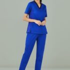Person in blue scrubs and white sneakers stands smiling against a gray background.
