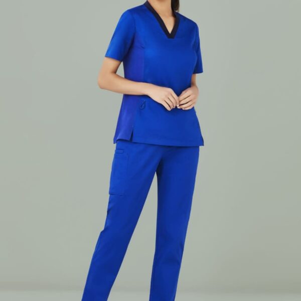 Person in blue scrubs and white sneakers stands smiling against a gray background.