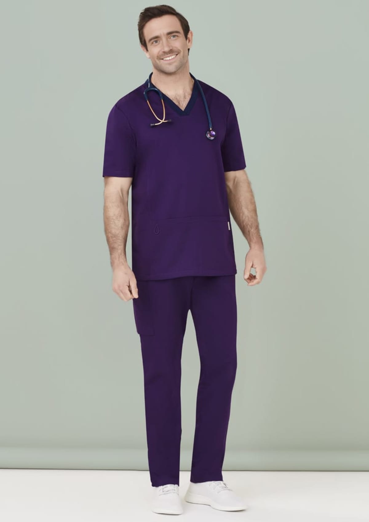 Person in purple medical scrubs with stethoscope, smiling against a gray background.