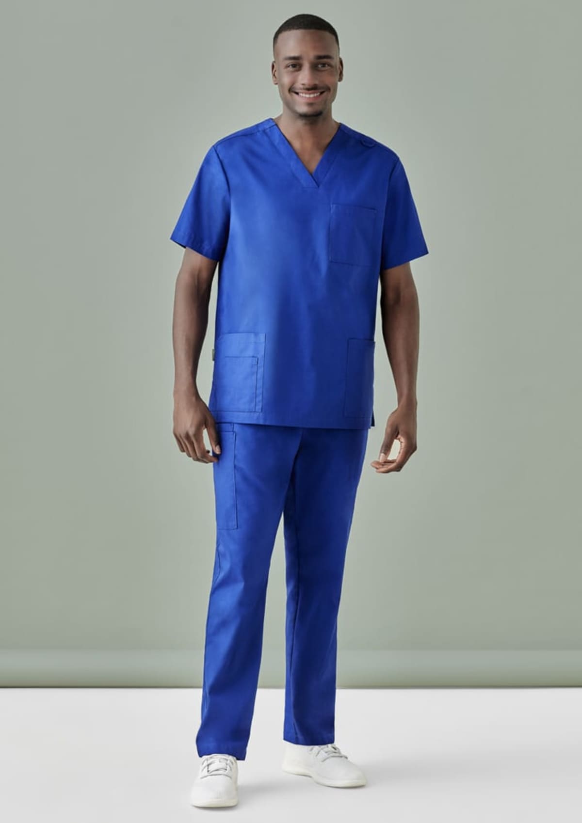 Person in blue medical scrubs and white sneakers stands against a plain backdrop.