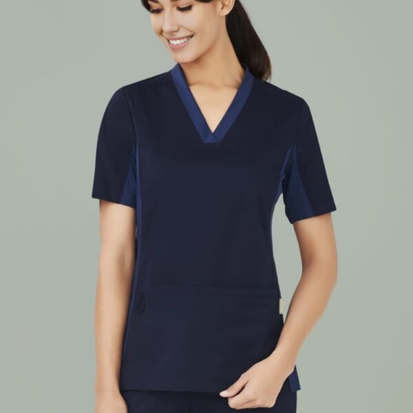 Woman in navy scrubs smiling, stands against a light gray background.