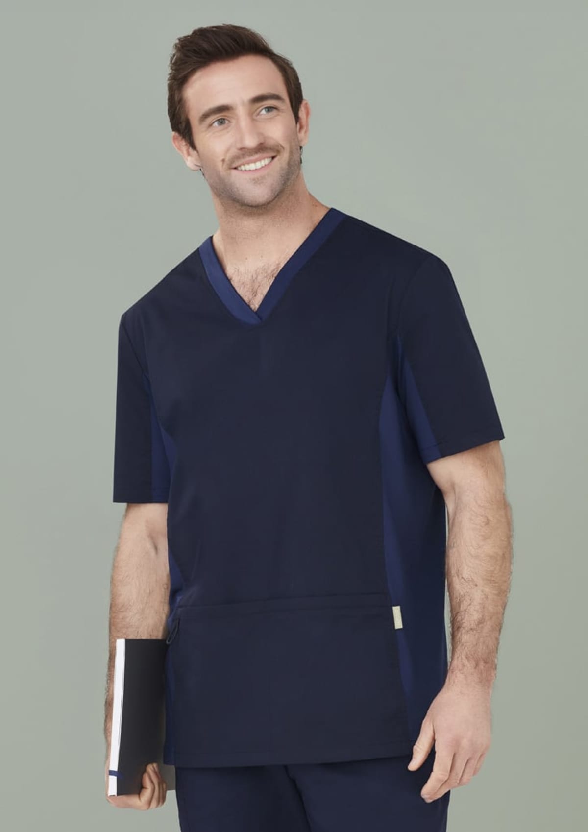 Smiling person in navy scrubs holding a notebook against a light green background.