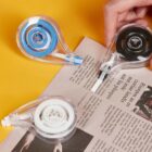 Hand using correction tape rollers on an open newspaper.