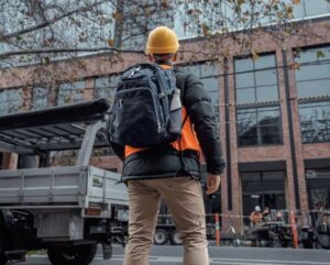 Construction worker with a custom hard hat and backpack stands by a building and truck.