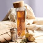 Glass bottle with tea, bamboo lid, and tea infuser beside ginger and herbs.