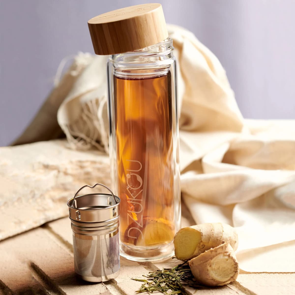 Glass bottle with tea, bamboo lid, and tea infuser beside ginger and herbs.