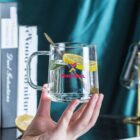A hand holds a glass mug of lemon water with a spoon, books and a lemon in the background.