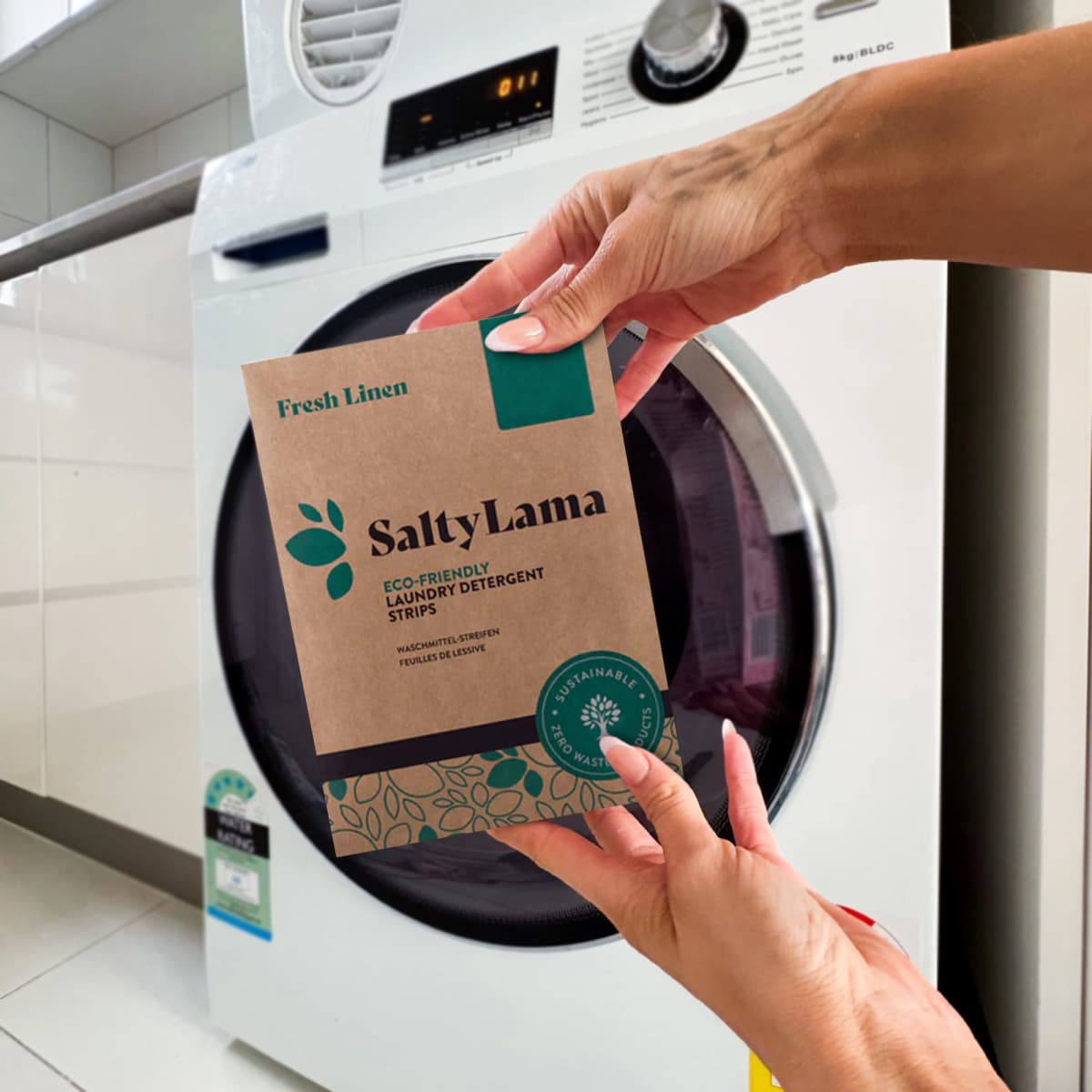 Hands holding SaltyLama laundry detergent strips in front of a washing machine.