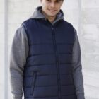 Person in a navy vest and gray hoodie, wearing a blue cap, stands against a concrete wall.