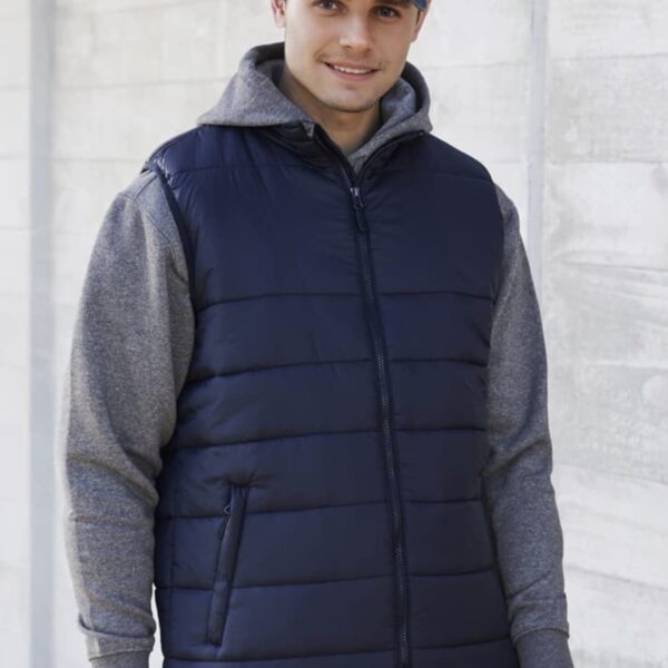 Person in a navy vest and gray hoodie smiles, wearing a denim cap, against a concrete wall.