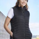 Woman in black puffer vest and beanie, standing outside with a blue sky background.