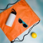 Orange drawstring bag with a white bottle, blue sunglasses, and a yellow ball on a light blue surface.