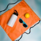 Orange drawstring bag, water bottle, blue sunglasses, and yellow ball on a light blue background.