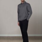 Man in a gray long-sleeve shirt and black pants stands smiling against a gray background.