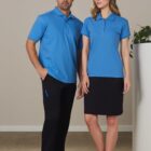 Two people wearing blue polos; one in pants and one in a skirt, standing indoors.