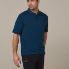 Man in a blue polo shirt and black shorts looking to the side, standing against a beige background.