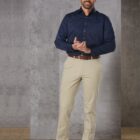 Man in navy shirt and beige pants standing against a gray wall.