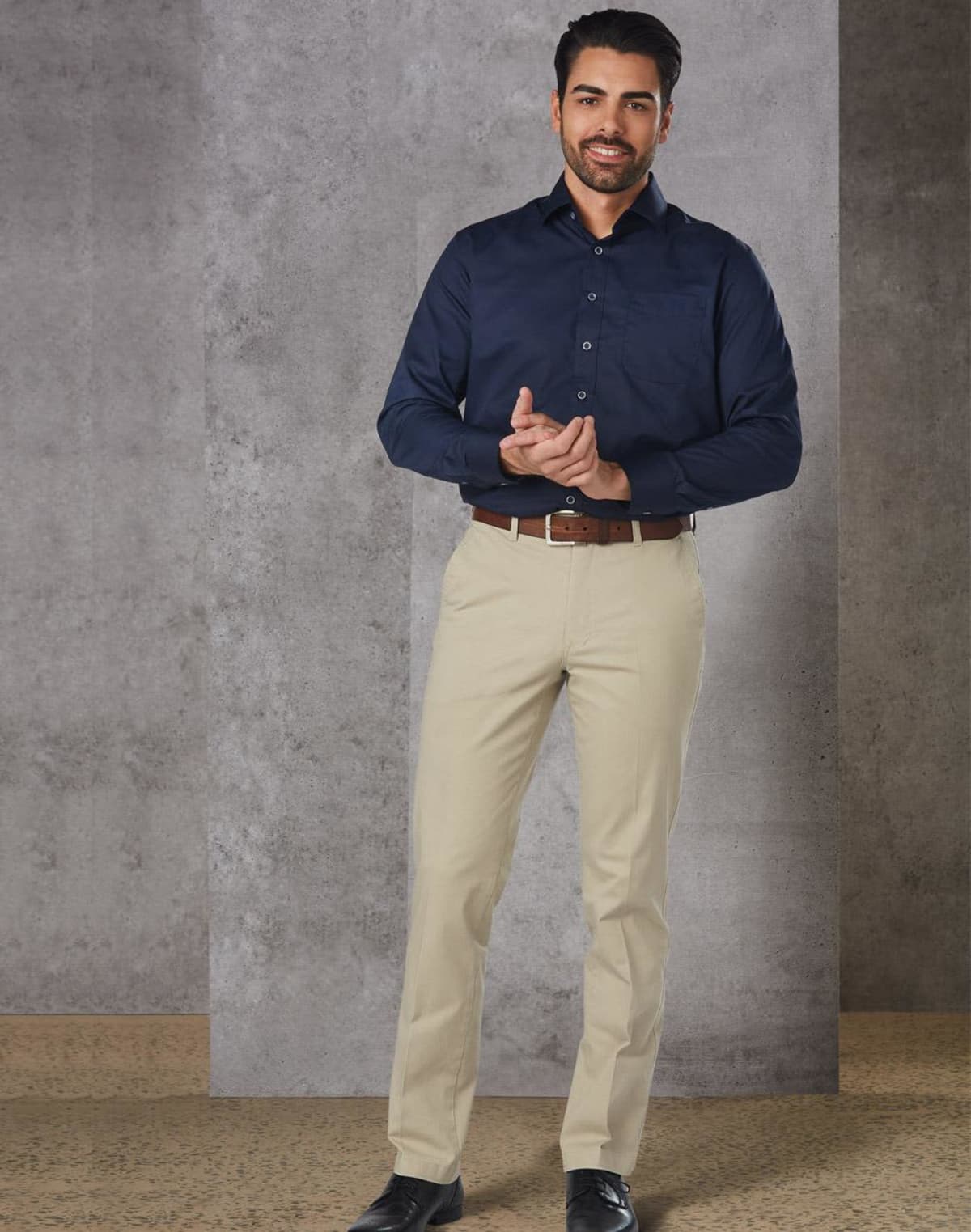Man in navy shirt and beige pants standing against a gray wall.